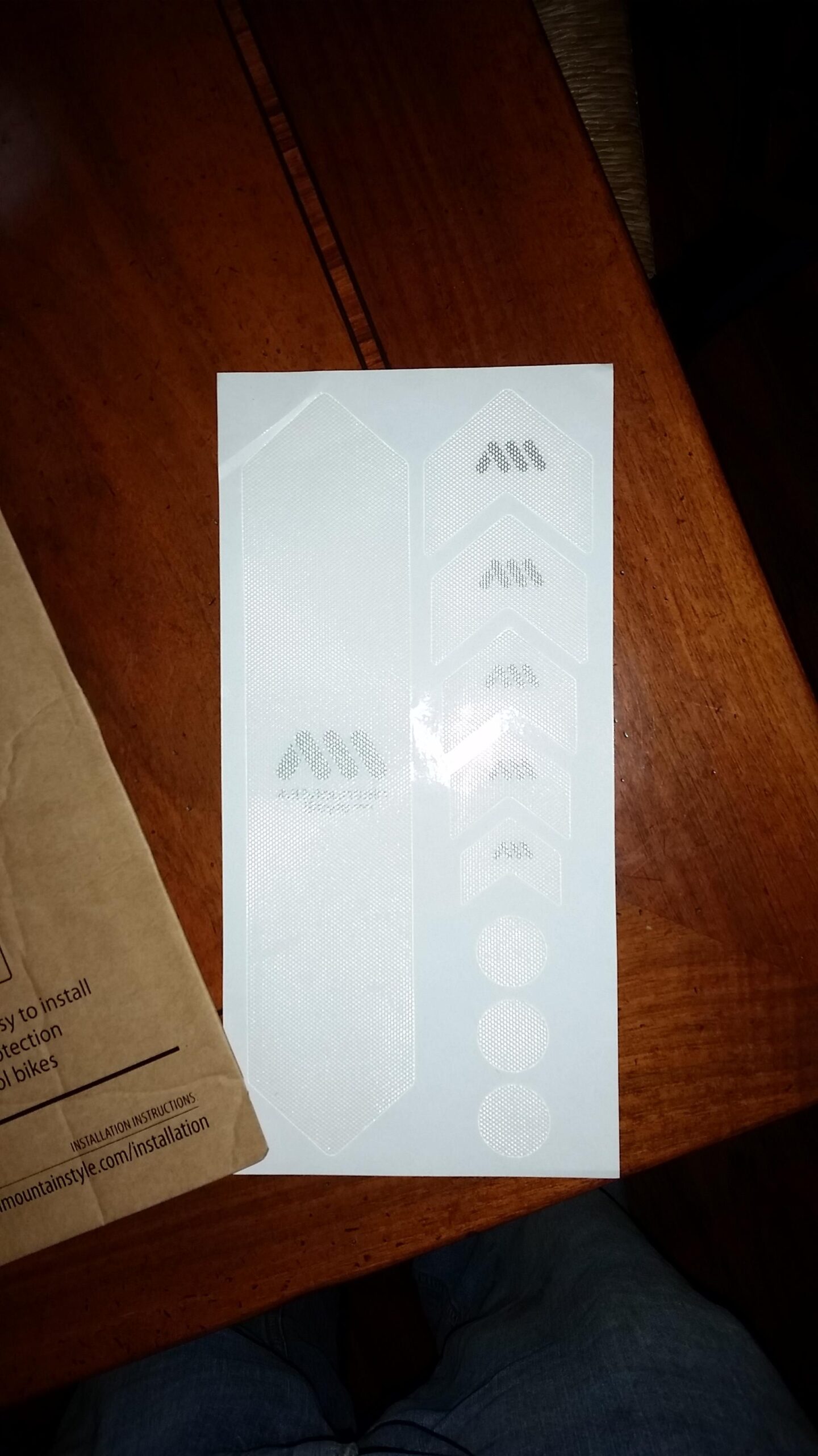 Motobecane NightTrain Bullet: Clear adhesive protective stickers arranged on a sheet, featuring various shapes such as hexagons, arrows, and circles. The sheet is placed on a wooden surface, with a cardboard package partially visible in the background, indicating the product's use for installation on bikes.