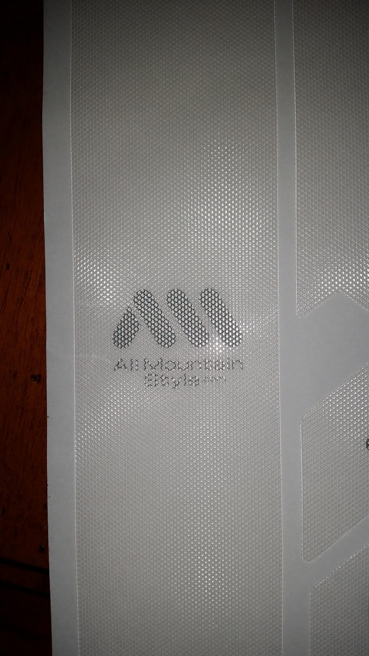 Motobecane NightTrain Bullet: Close-up view of a textured, transparent film with a logo featuring three stylized peaks, accompanied by the text "All Mountain Style." The logo is printed on a white background, and the film has a characteristic dotted pattern.