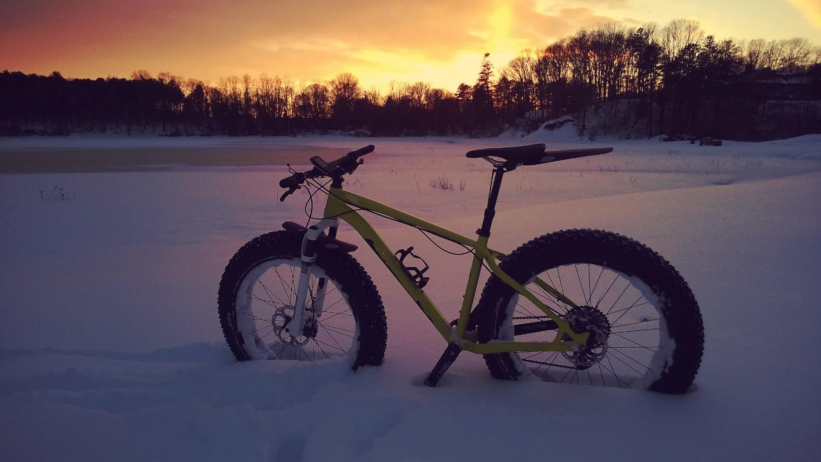 Specialized Fatboy Pro: A bright yellow fat bike stands in deep snow with a picturesque sunset in the background, featuring hues of orange and purple. The scene captures a winter landscape with trees silhouetted against the colorful sky.