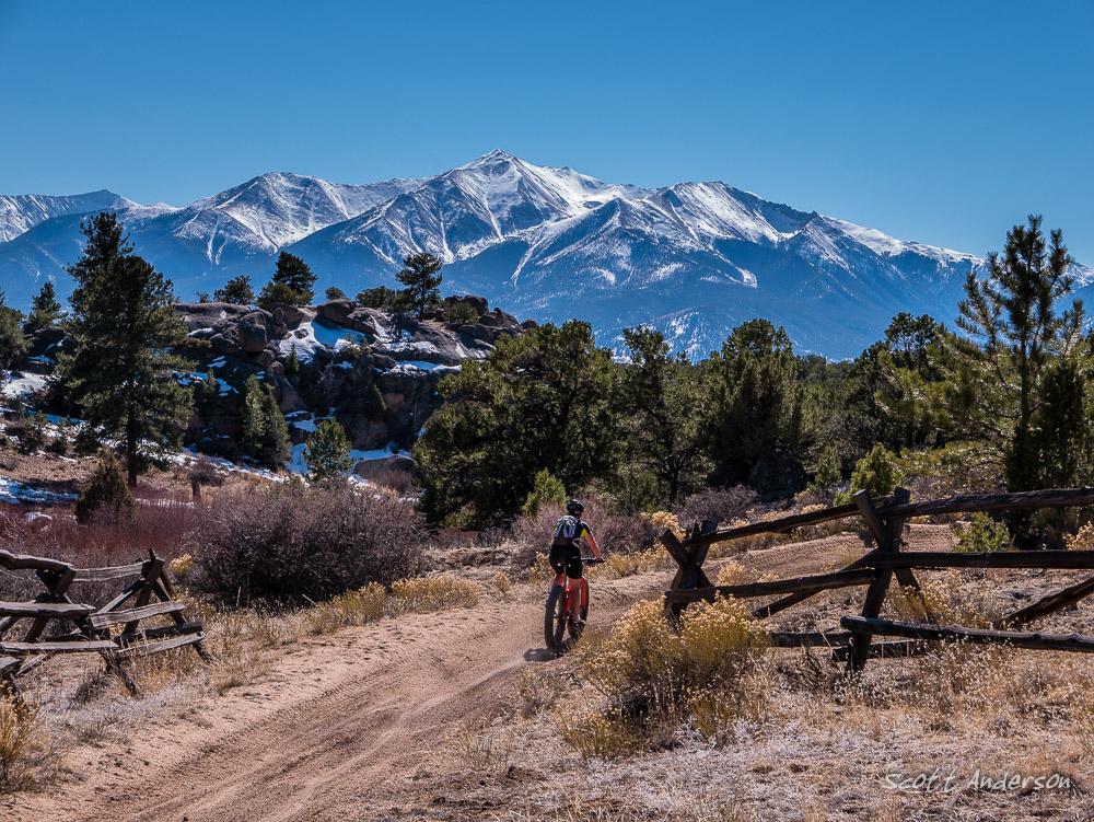 A mountain biker rides along a dirt trail surrounded by lush vegetation, with snow-capped mountains in the background under a clear blue sky. Wooden fences line the path, adding to the scenic outdoor landscape. Trail #1415 mountain bike trail.