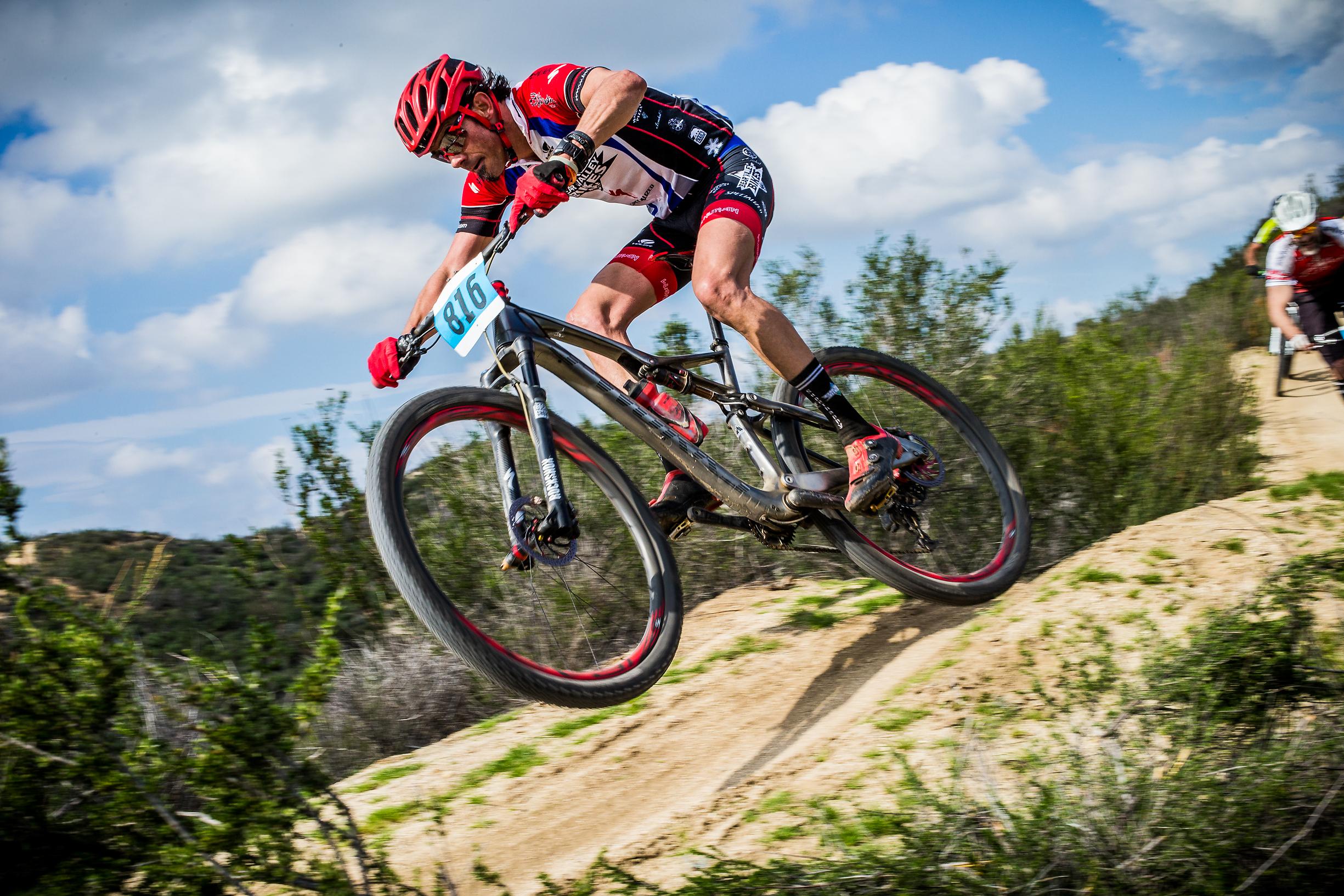 Specialized S-Works Epic WC: A mountain biker in a red jersey and helmet performs a jump over a dirt ramp, surrounded by greenery and under a partly cloudy sky. Another cyclist can be seen in the background, also riding on the trail. The scene captures the excitement of mountain biking and the athlete's dynamic movement in mid-air.