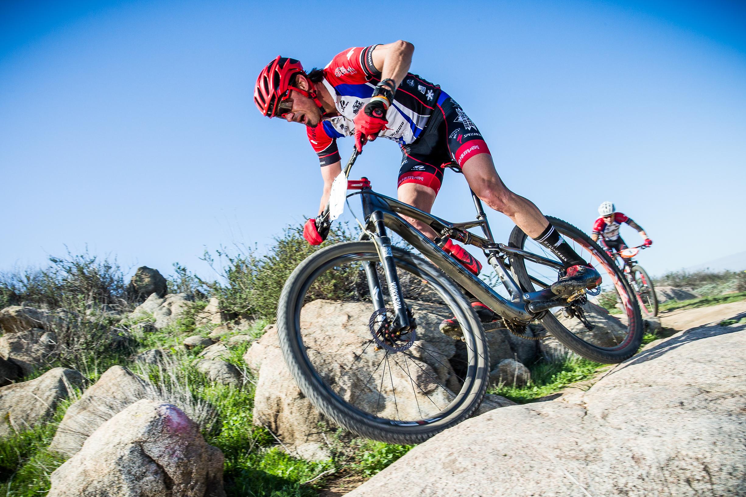 Specialized S-Works Epic WC: A mountain biker in a red helmet and cycling gear navigates over rocky terrain during a race, showcasing skill and focus. In the background, another cyclist follows closely behind, set against a clear blue sky and green landscape.