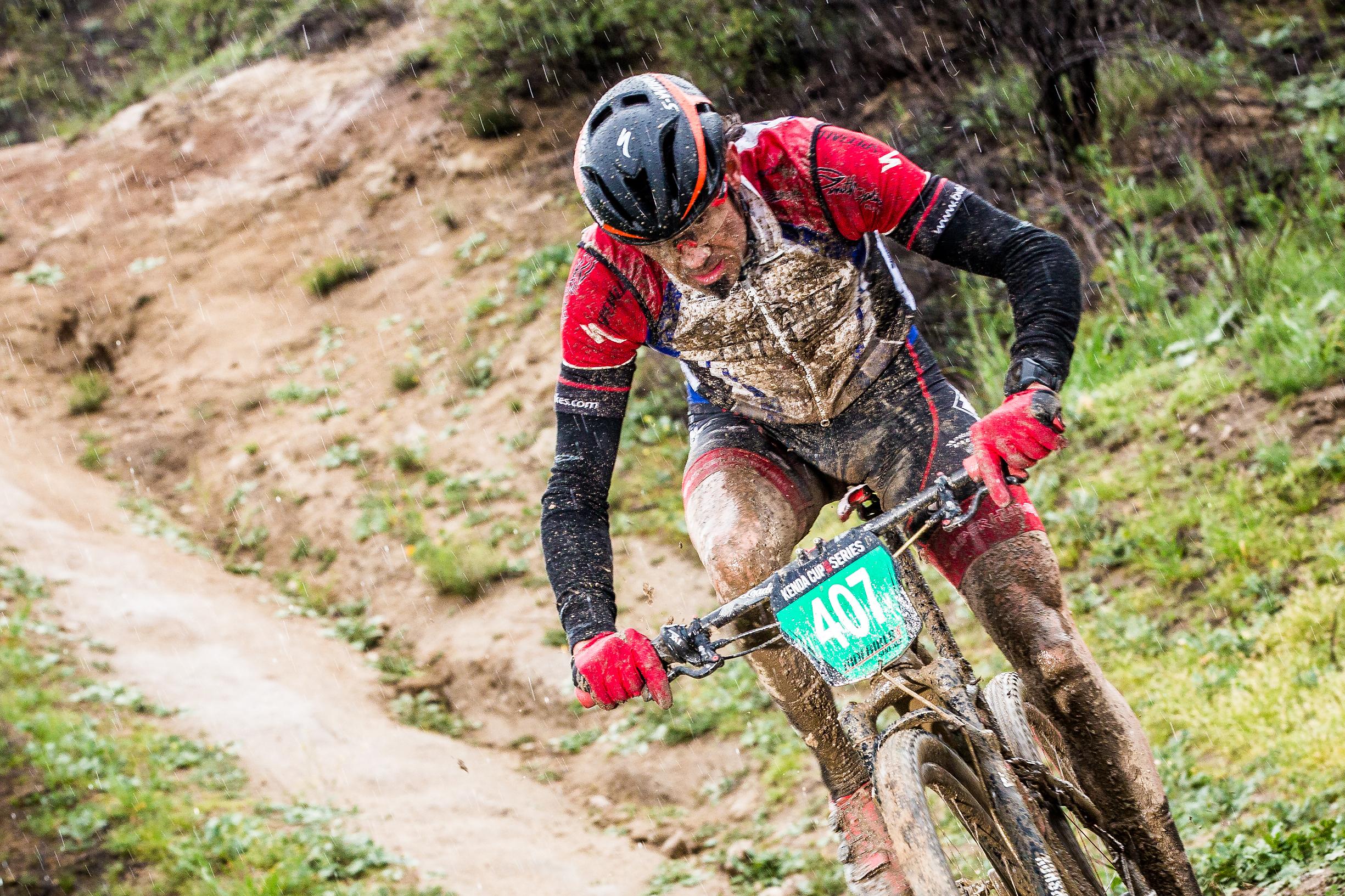 Specialized S-Works Epic WC: A mountain biker wearing a red and black jersey and helmet rides along a muddy trail during a rainstorm, covered in mud and exerting effort as water droplets fall around him. The bike features a race number on the side, indicating participation in a competition.