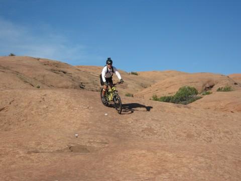 Giant Yukon: A person riding a mountain bike on a rocky terrain under a clear blue sky. The landscape features smooth, reddish-brown rock formations with some sparse greenery.