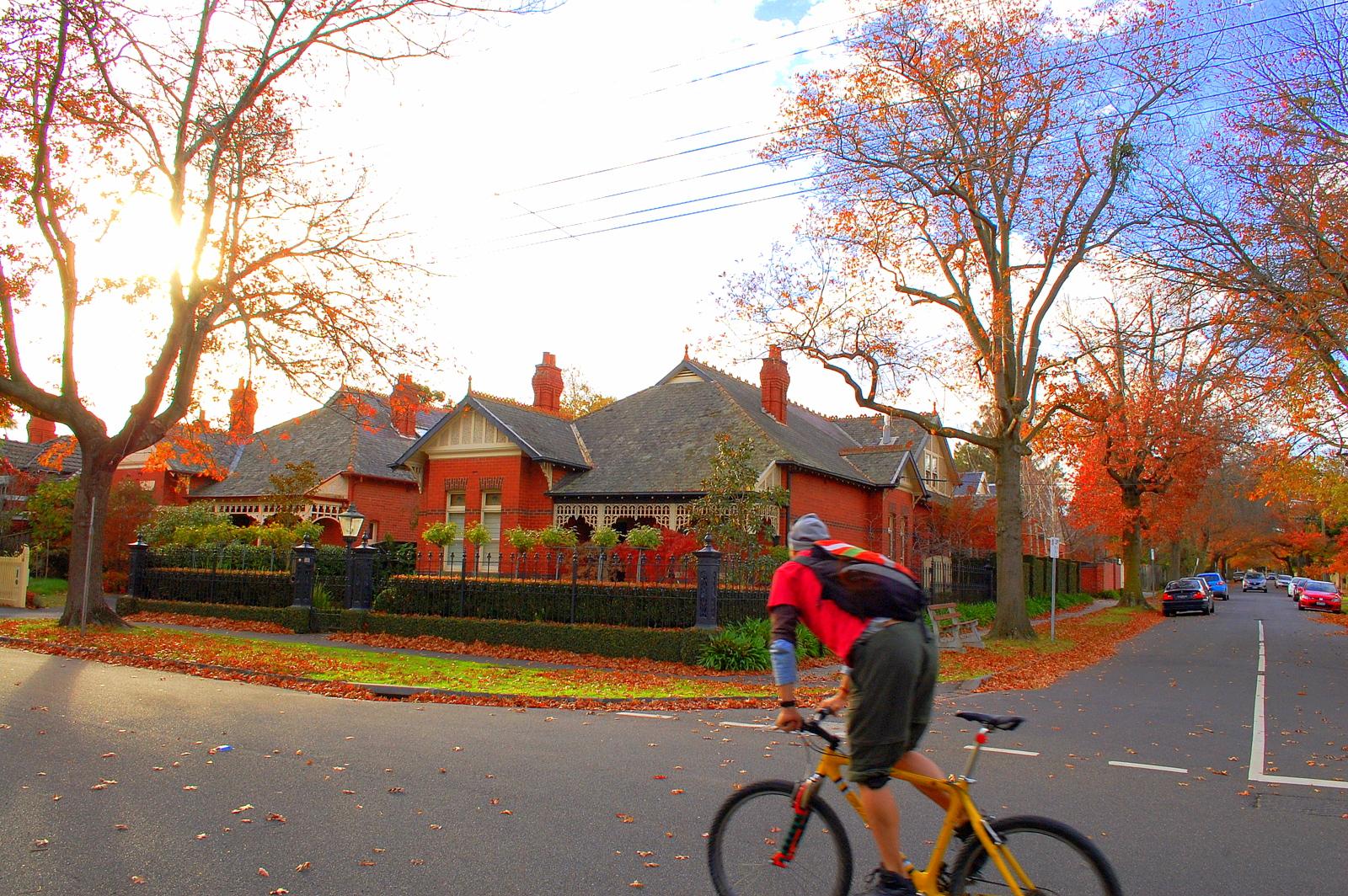 Giant MCM ONE Carbon Fibre Mountain Bike: A cyclist in a red shirt rides past a charming brick house surrounded by autumn foliage, with colorful leaves scattered on the ground. The scene captures a bright autumn day as sunlight filters through the trees, highlighting the warm colors of the season.
