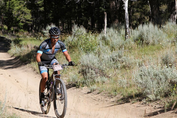 A mountain biker riding on a dirt trail surrounded by greenery and trees. The cyclist is wearing a helmet, sunglasses, and a cycling jersey with the number 102 displayed on the bike.
