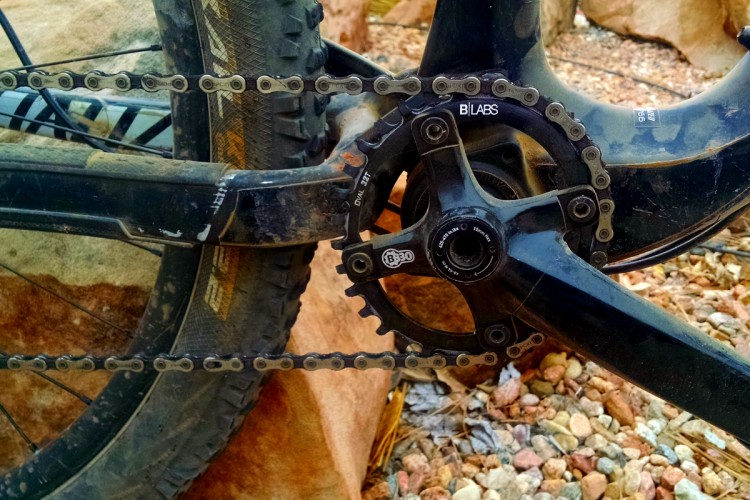 Close-up view of a mountain bike's drivetrain, featuring a black crankset, chainring, and chain. The image shows a dirty bike with a rugged tire set against a rocky terrain.