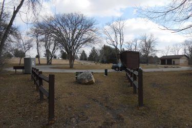 A park scene featuring a dirt path winding through an open area with bare trees. A wooden fence runs through the foreground, while a picnic table and a large rock are visible. In the background, there is a portable restroom, a parked vehicle, and an additional building. The sky is partly cloudy, suggesting a cool day. Rockport Park MTB mountain bike trail.
