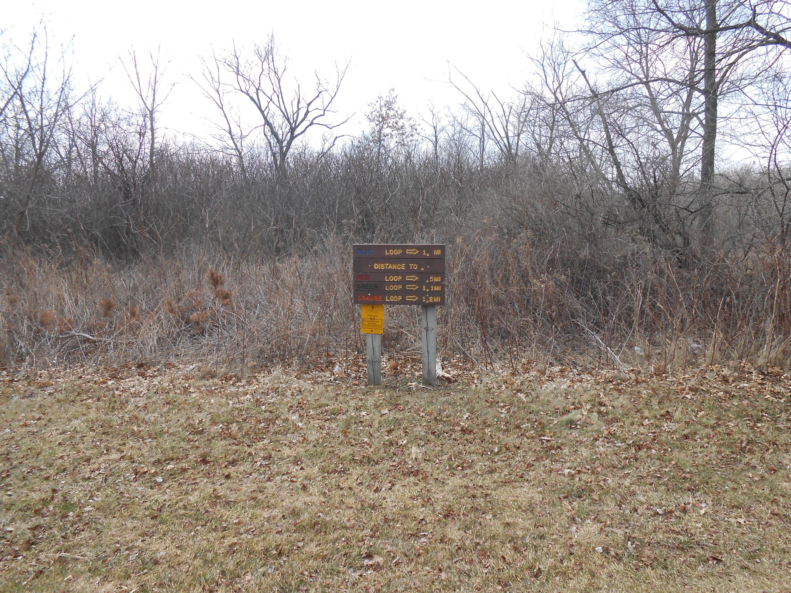 Signpost with multiple trails indicated, showing loop distances in miles: blue loop (1 mile), red loop (0.5 miles), green loop (1 mile), and orange loop (1.2 miles). The background features sparse trees and brush in a grassy area during overcast weather. Rockport Park MTB mountain bike trail.