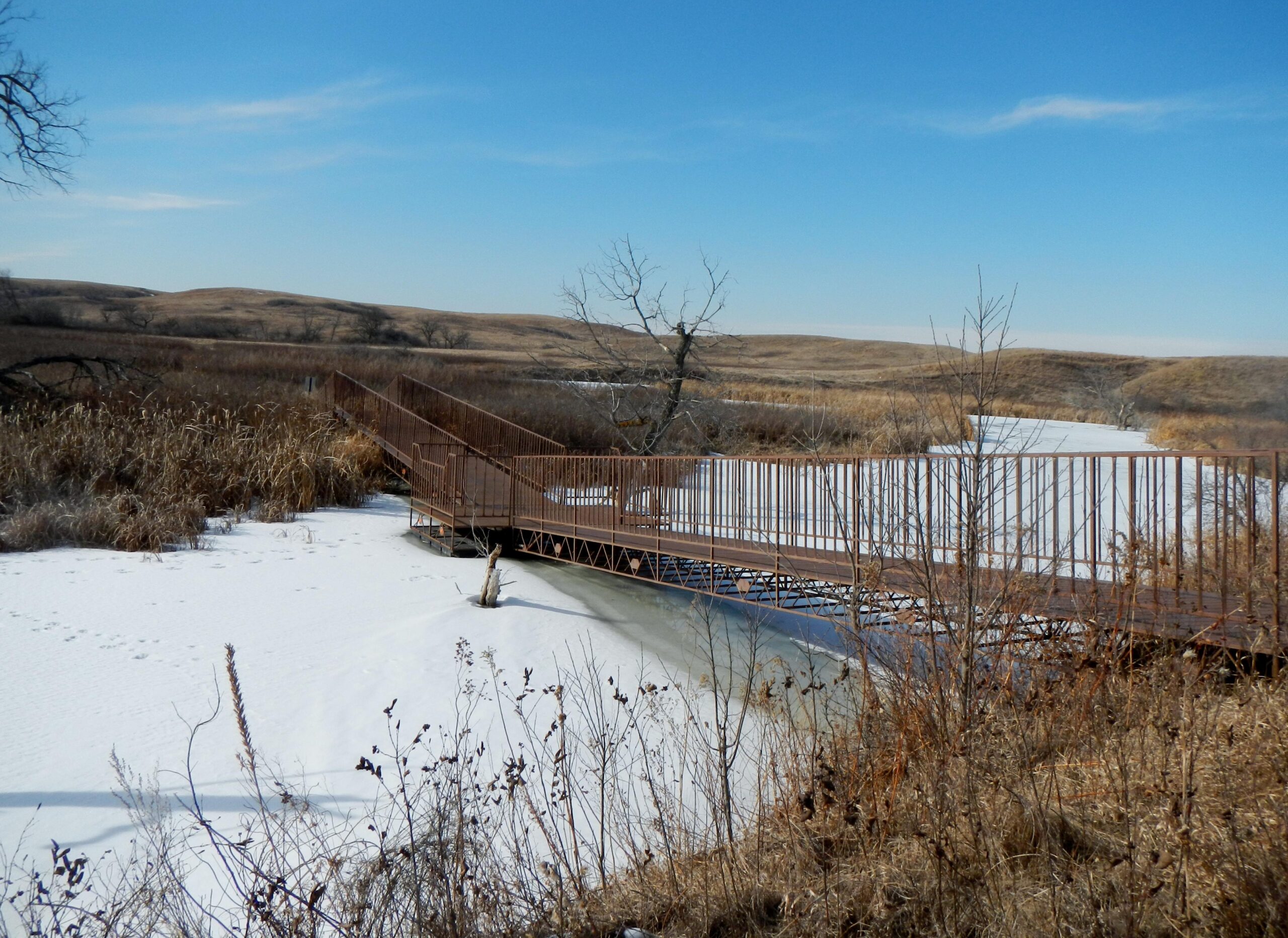 A winter landscape featuring a wooden bridge over a partially frozen waterway surrounded by snow-covered ground and dry grasses. In the background, rolling hills are visible under a clear blue sky. Harmon mountain bike trail.