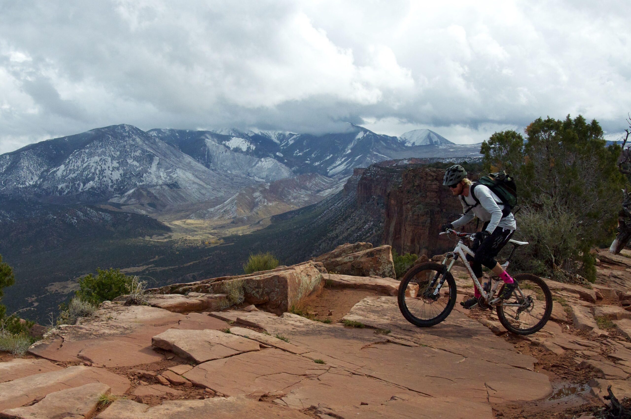 A mountain biker navigating a rocky trail with mountainous terrain and cloudy skies in the background. The landscape features snow-capped peaks and rugged cliffs, showcasing the beauty of nature and outdoor adventure. Porcupine Rim mountain bike trail.