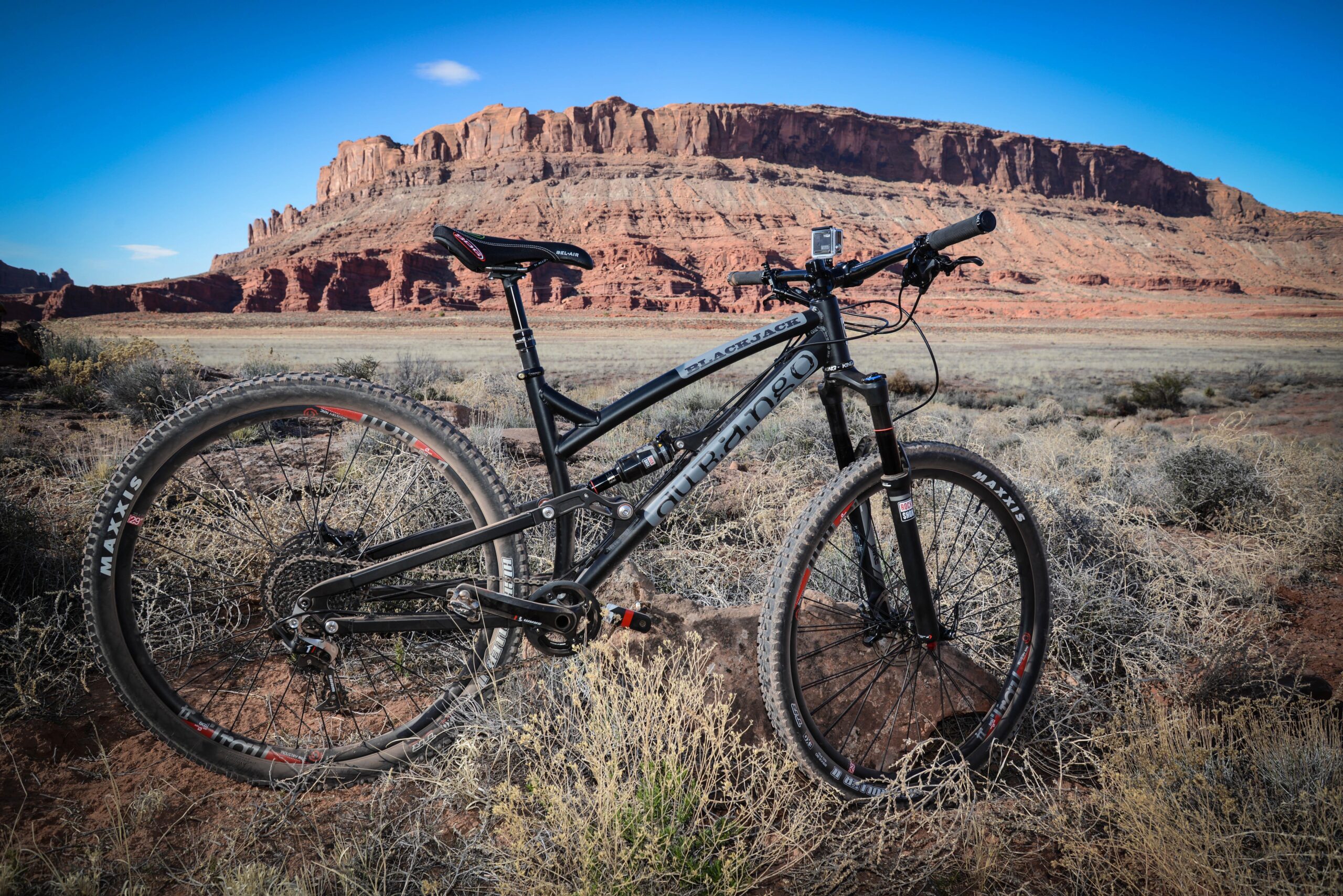 Durango Bike Company Blackjack: A black mountain bike is positioned on rocky terrain in a desert landscape, with red rock formations in the background under a clear blue sky. The scene showcases the bike's details, including its tires and frame, highlighting an adventurous outdoor setting.