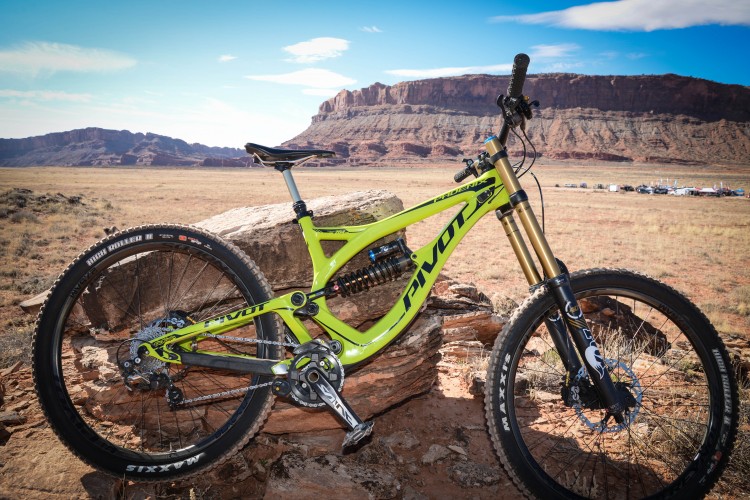 A bright green Pivot mountain bike is positioned on a rocky outcrop in a vast outdoor landscape. In the background, red rock formations and a clear blue sky are visible, creating an adventurous setting. The bike features prominent components, including a front suspension fork and wide tires, indicating its suitability for rugged terrain.