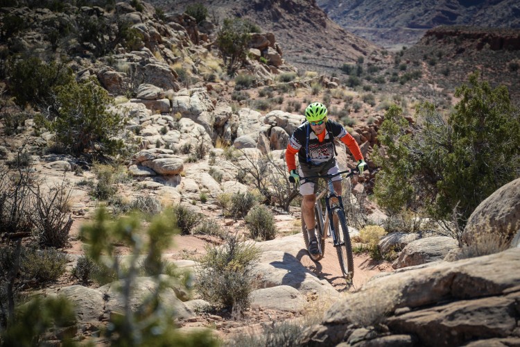 Mountain biker navigating rocky terrain in a desert landscape, wearing a green helmet and orange sleeves, surrounded by shrubs and rocky formations.