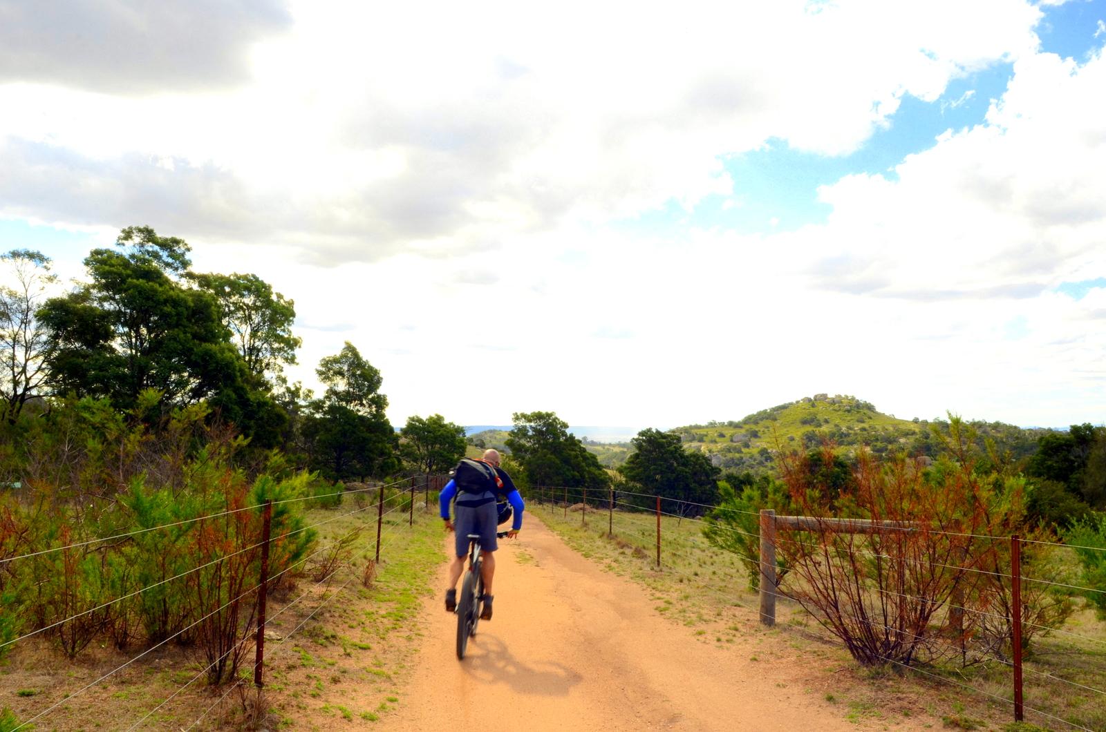 A person riding a mountain bike on a dirt trail, surrounded by lush greenery and trees. The path is bordered by a wire fence, with hills visible in the background under a partly cloudy sky. You Yangs Regional Park mountain bike trail.