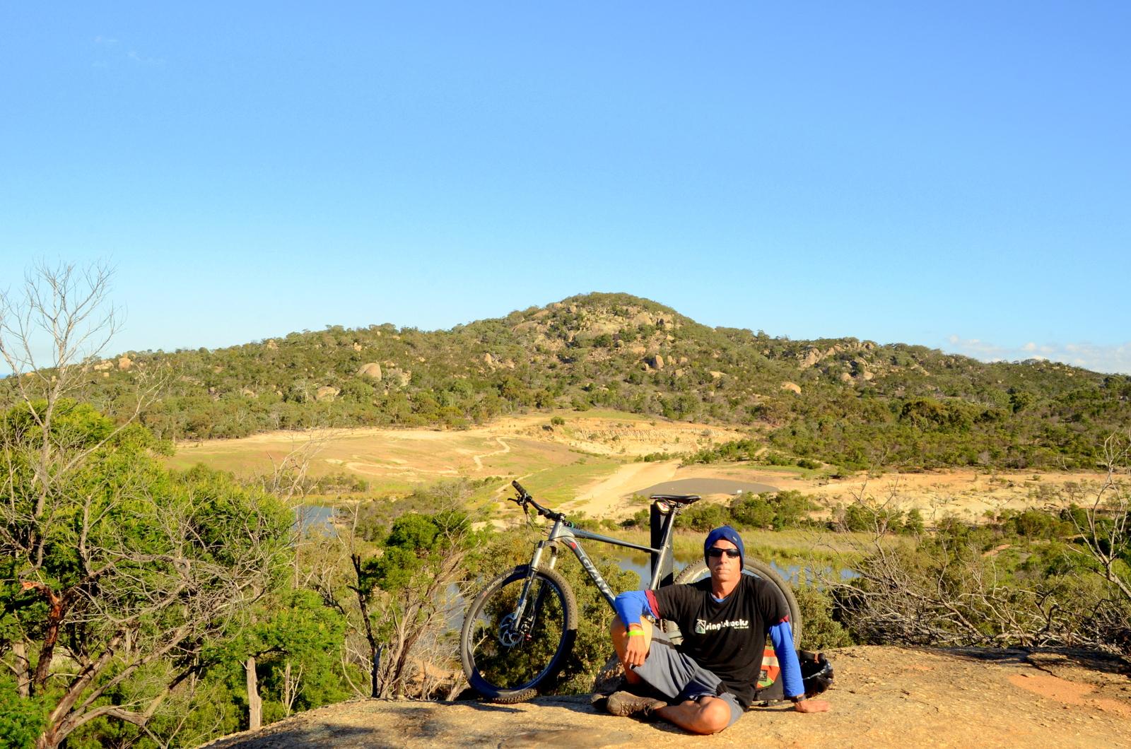 A person sitting on a rocky outcrop with a mountain bike nearby, surrounded by a scenic landscape featuring hills, trees, and a clear blue sky. The individual is wearing casual outdoor clothing and sunglasses, appearing relaxed in a natural setting. You Yangs Regional Park mountain bike trail.