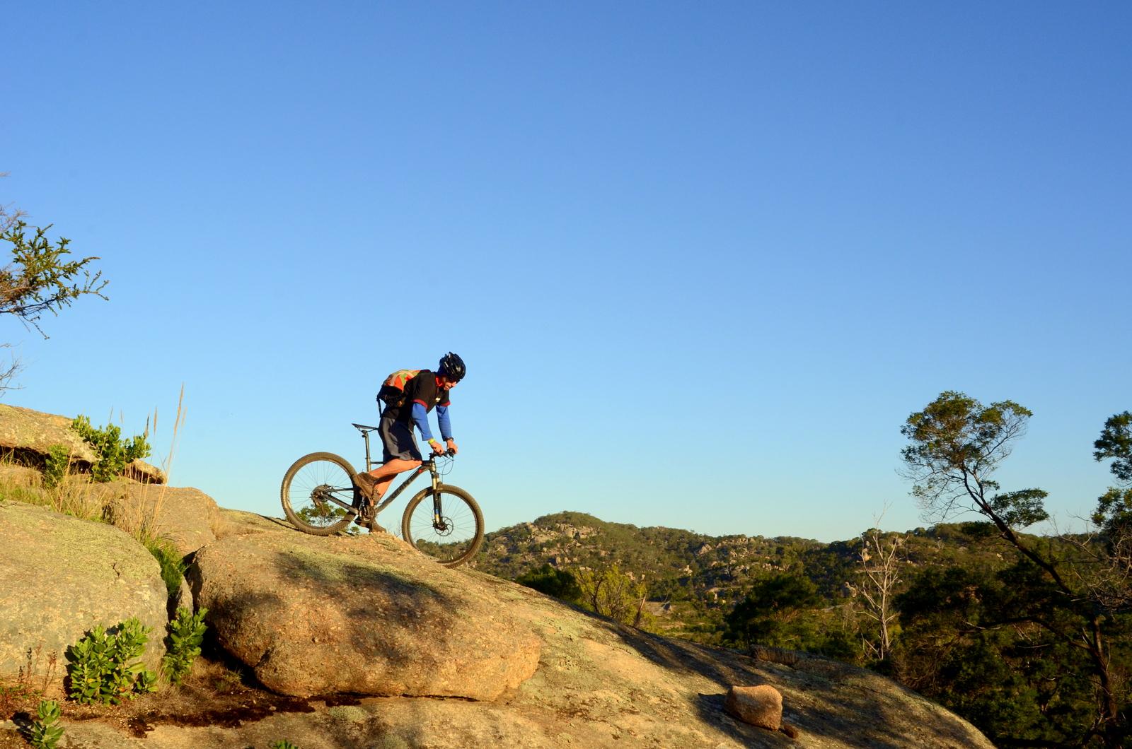 A mountain biker navigating rocky terrain against a clear blue sky, surrounded by hills and sparse vegetation. The cyclist is wearing a helmet and riding a mountain bike, focusing on balancing on large boulders. You Yangs Regional Park mountain bike trail.