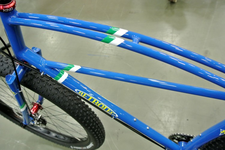 A close-up view of a blue bicycle frame showcasing unique curved top tubes and green and white accents. The rugged tires are visible in the foreground, and the background appears to be a smooth, light-colored flooring typical of an indoor space.