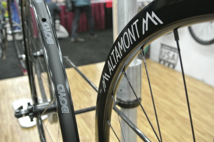 Close-up view of bicycle wheels featuring the Boyd and Altamont brands. The wheels have a sleek black finish, with visible spokes and logos prominently displayed. The background is softly blurred, suggesting an indoor setting, possibly a bike expo.