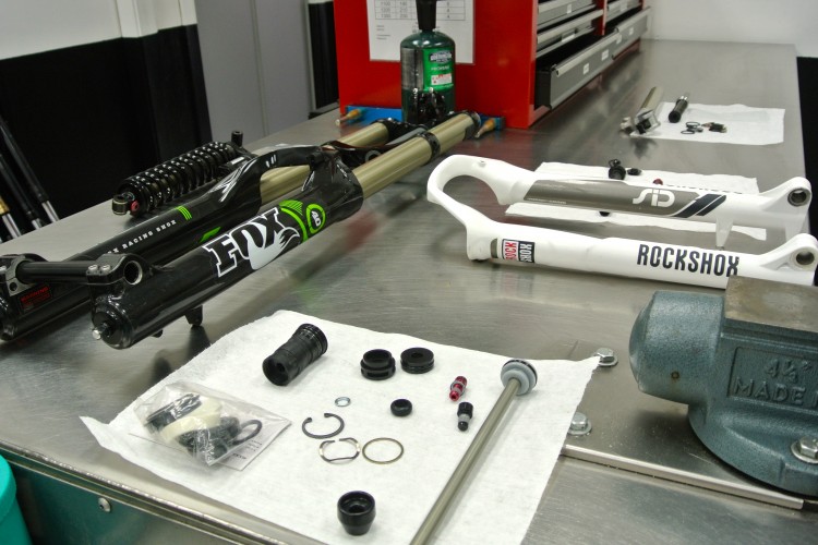 Collection of bicycle suspension components displayed on a workbench, including a disassembled FOX Racing shock and RockShox fork. Various small parts, tools, and a canister of lubricant are arranged on white paper towels, highlighting intricate details of the components.