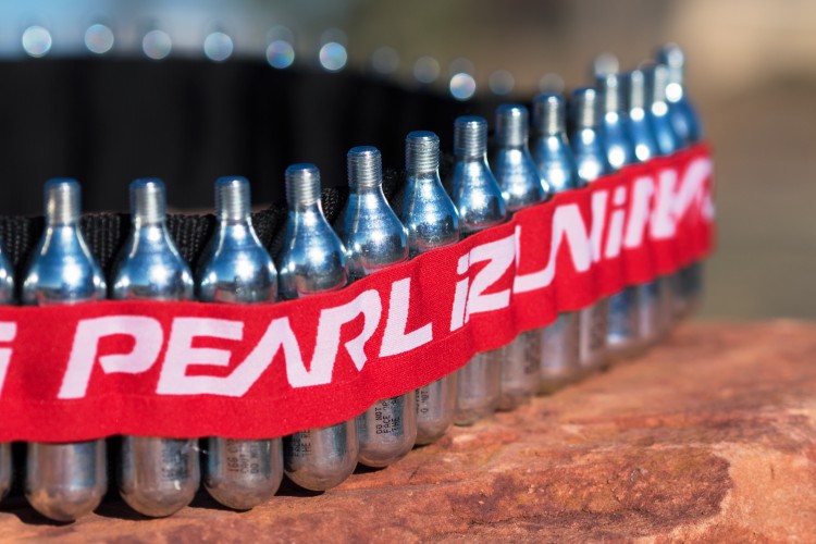 A close-up image of a red belt featuring several silver CO2 cartridges arranged in a row. The belt has a logo printed in white, and it appears to be resting on a rocky surface with a blurred background.