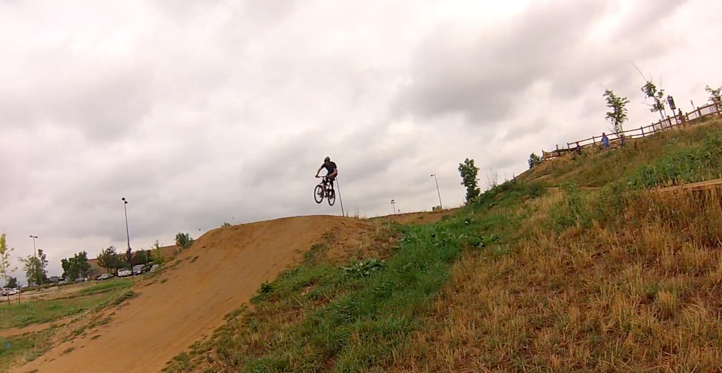 A cyclist performing a jump on a dirt ramp in a park, with overcast skies and green grass surrounding the area. Valmont Bike Park mountain bike trail.
