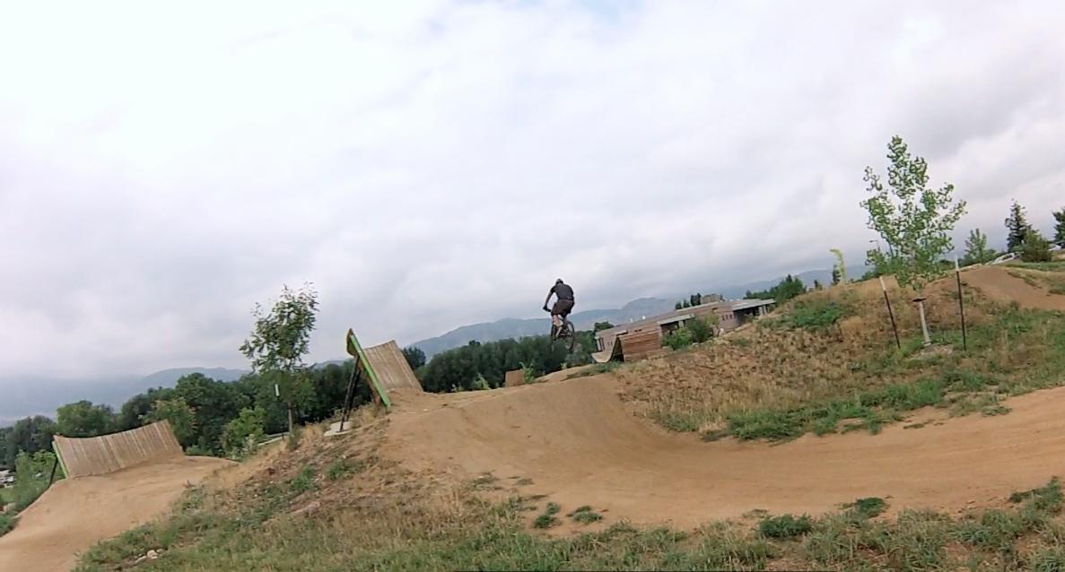 A cyclist performing a jump on a dirt track at a BMX park, surrounded by green trees and a cloudy sky in the background. Valmont Bike Park mountain bike trail.