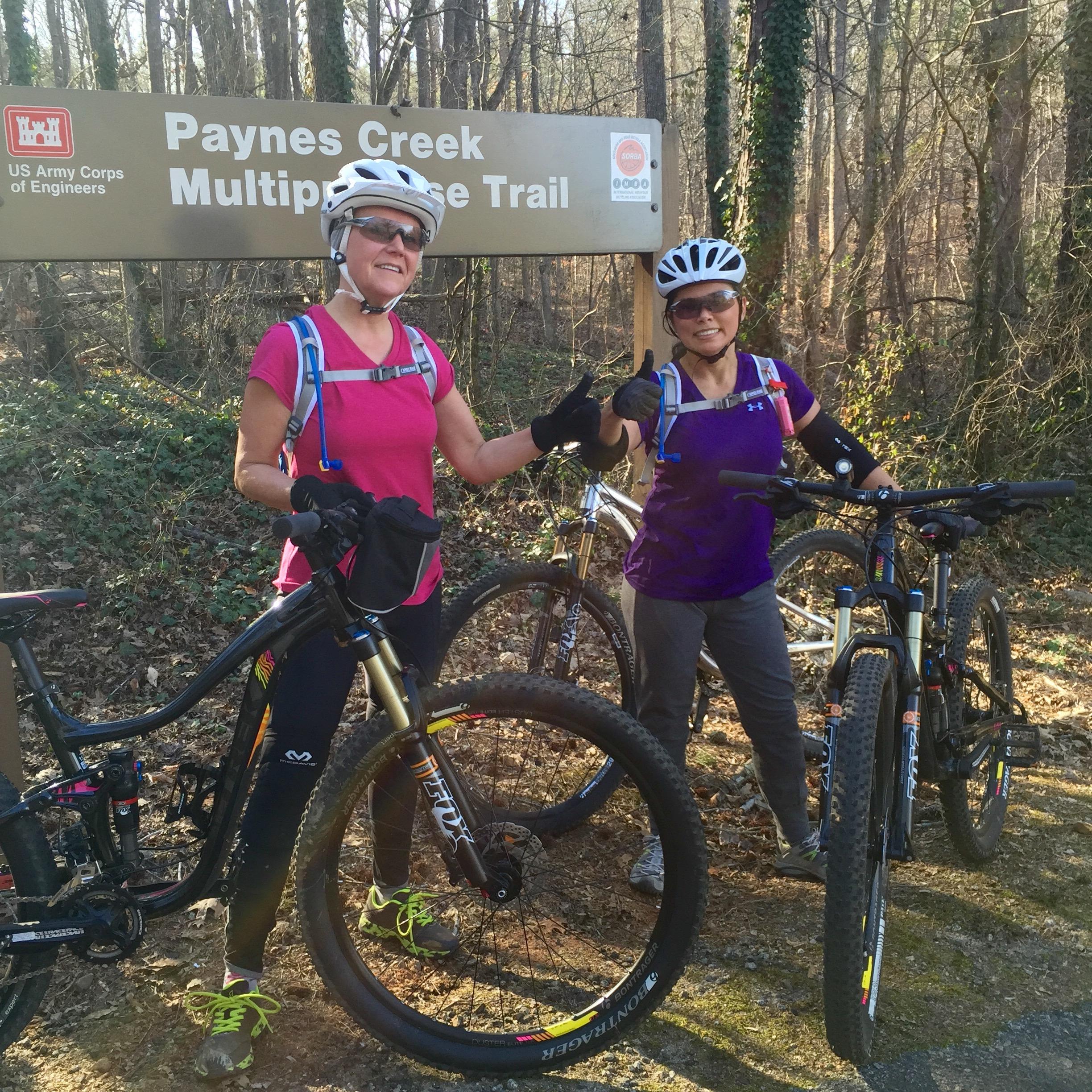 Two female mountain bikers are standing in front of a sign that reads "Paynes Creek Multipurpose Trail." They are both wearing helmets and biking attire, with one giving a thumbs-up gesture. Bicycles are positioned beside them, and the background features a wooded area. Paynes Creek mountain bike trail.
