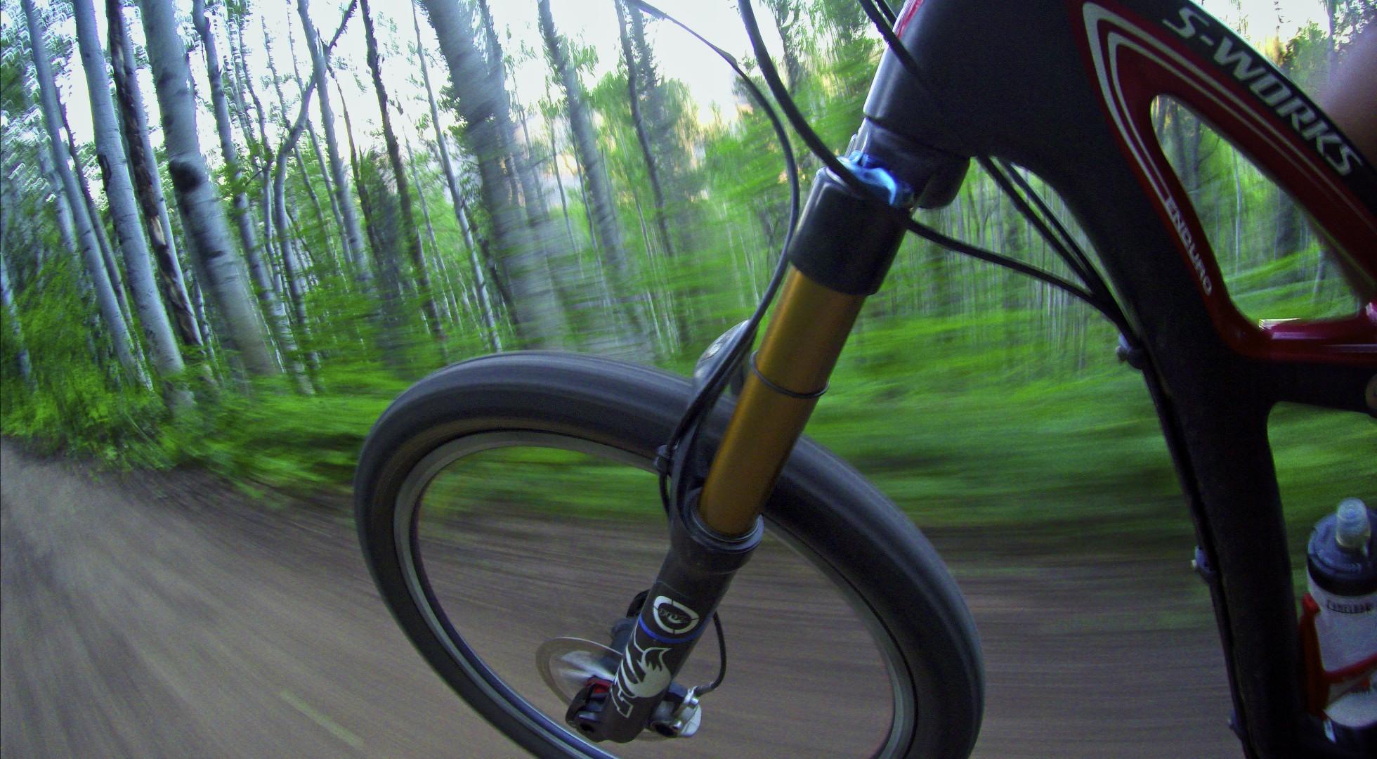 Close-up of a bicycle wheel in motion, on a dirt trail surrounded by green trees and foliage, suggesting a sense of speed and outdoor adventure. Beaver Creek Ski Resort mountain bike trail.