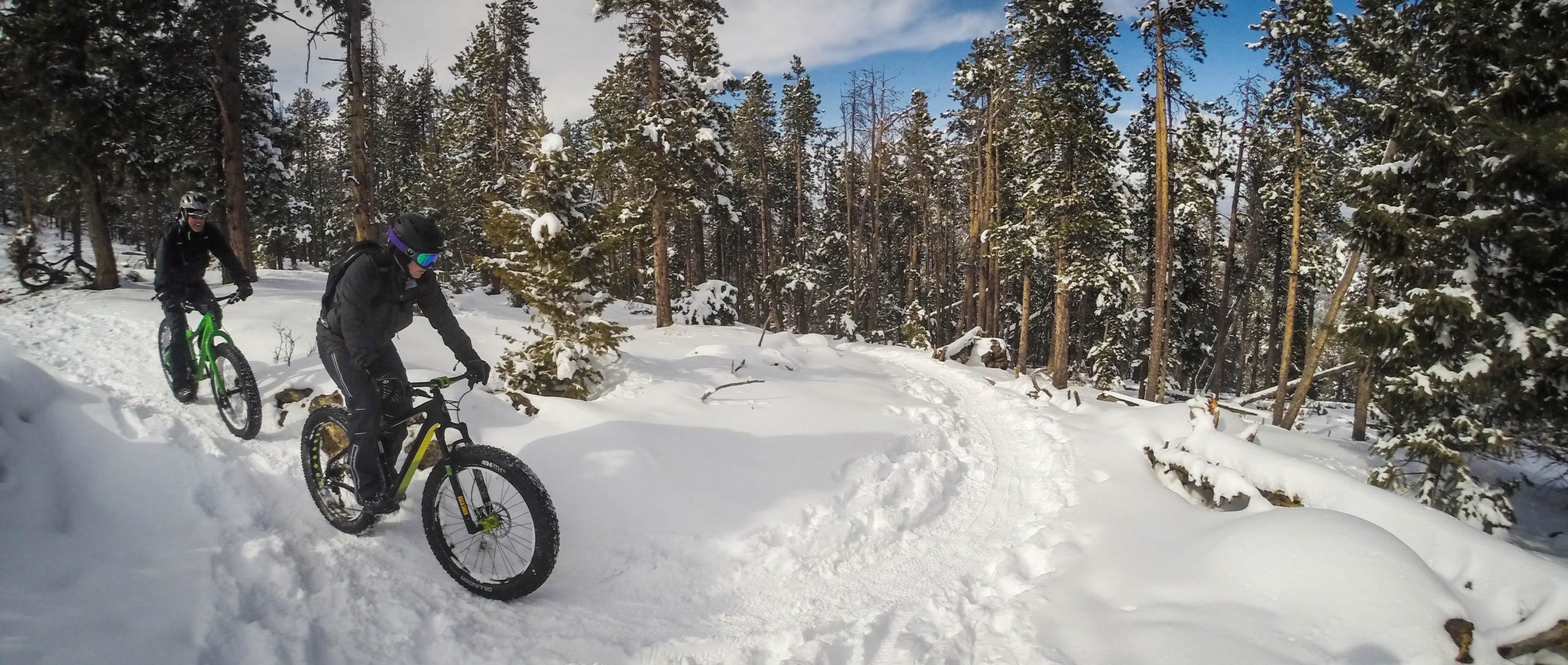 Two mountain bikers riding fat bikes along a snowy trail in a pine forest. The scene features snow-covered trees and a winding path, with one rider wearing a dark outfit and colorful goggles, and the other wearing a green fat bike. The atmosphere captures a winter outdoor adventure. 3 Sisters / Alderfer mountain bike trail.