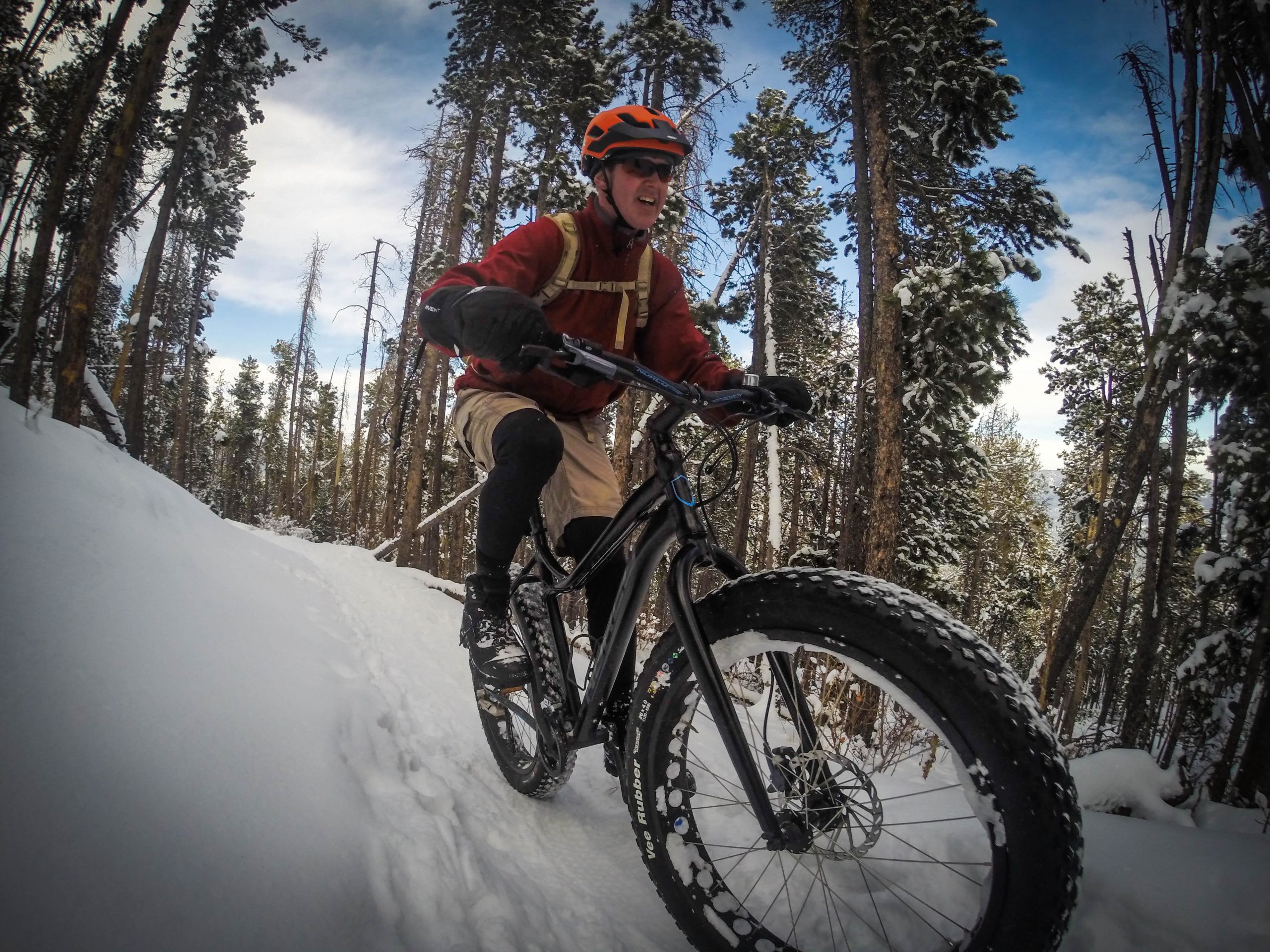A cyclist wearing a red jacket, black gloves, and an orange helmet rides a fat bike through snowy terrain surrounded by tall pine trees. The trail is partly covered in snow, and the sky is partly cloudy. The cyclist appears enthusiastic as they navigate the snowy path. 3 Sisters / Alderfer mountain bike trail.