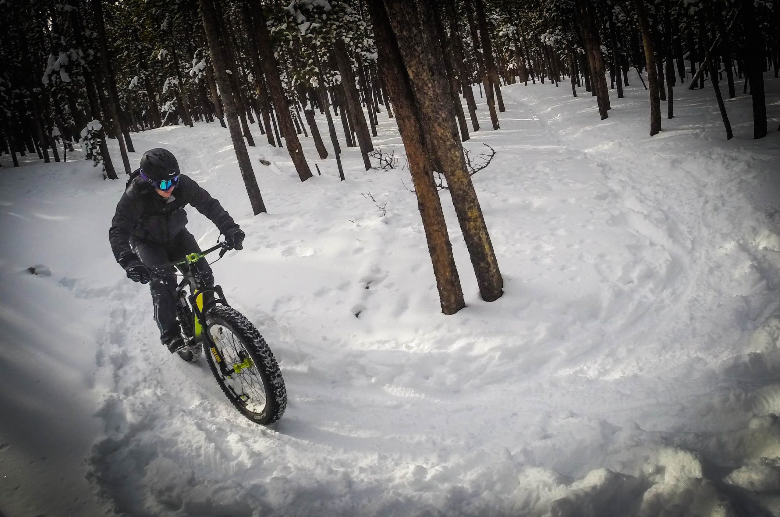 A person riding a fat bike through snow-covered trails in a forest. The cyclist is wearing a helmet and goggles, navigating a snowy path surrounded by tall trees. 3 Sisters / Alderfer mountain bike trail.