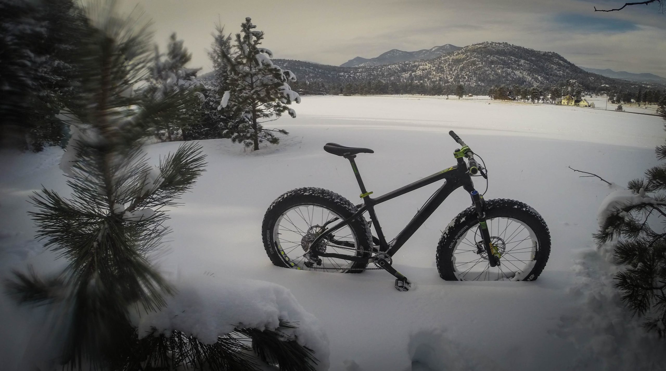 A black fat bike with thick tires stands on a snowy landscape surrounded by pine trees, with mountains in the background. The scene depicts a serene winter setting with fresh, untouched snow and a cloudy sky. 3 Sisters / Alderfer mountain bike trail.