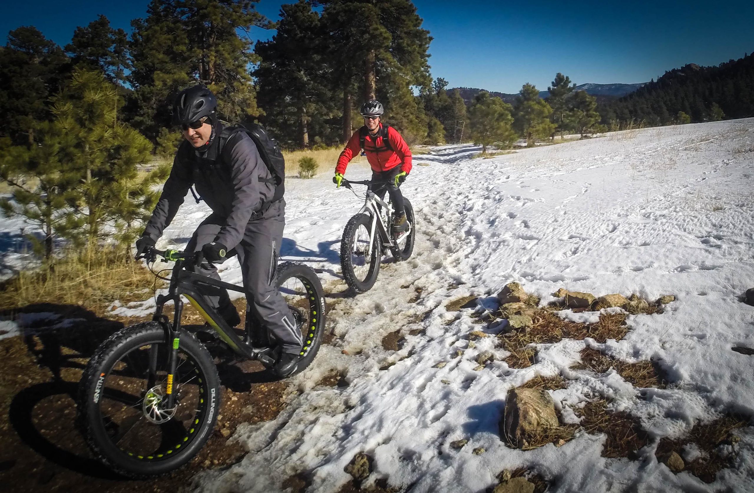 Two cyclists riding fat bikes on a snowy trail surrounded by trees. One cyclist is wearing a grey outfit and a helmet, while the other is dressed in a red jacket and black gear. The terrain is a mix of snow and rocky patches, indicating winter conditions. The background features a clear blue sky and a forested landscape in the distance. 3 Sisters / Alderfer mountain bike trail.