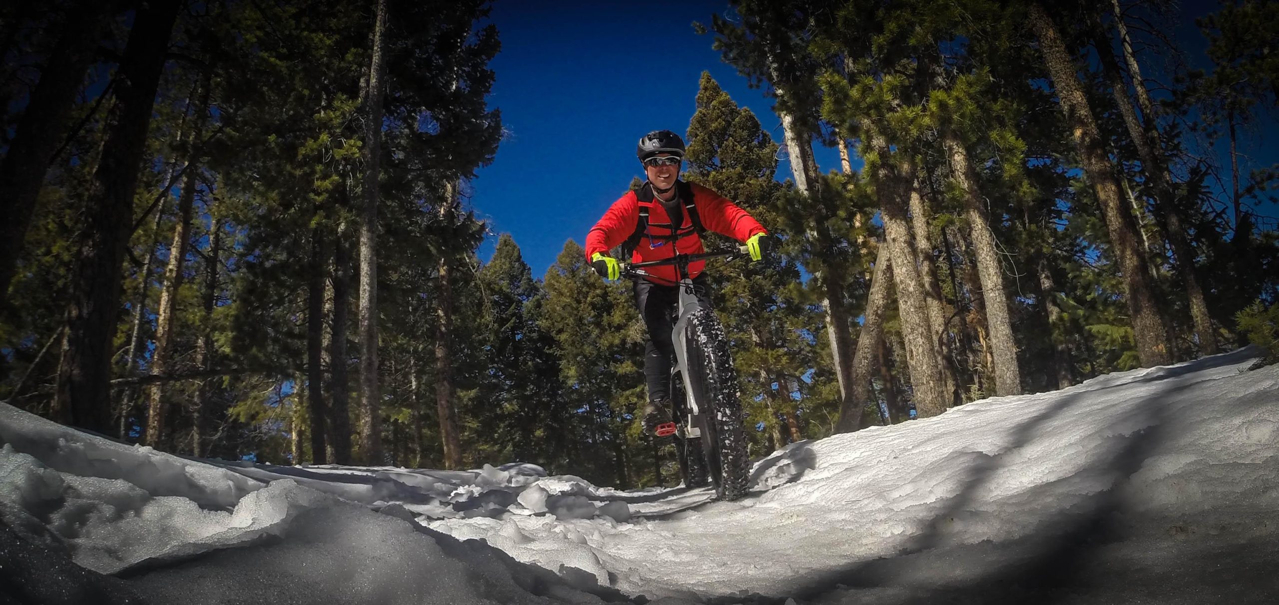 A person riding a mountain bike on snow-covered terrain in a forest, wearing a red jacket, black pants, and a helmet, with tall trees and a clear blue sky in the background. 3 Sisters / Alderfer mountain bike trail.
