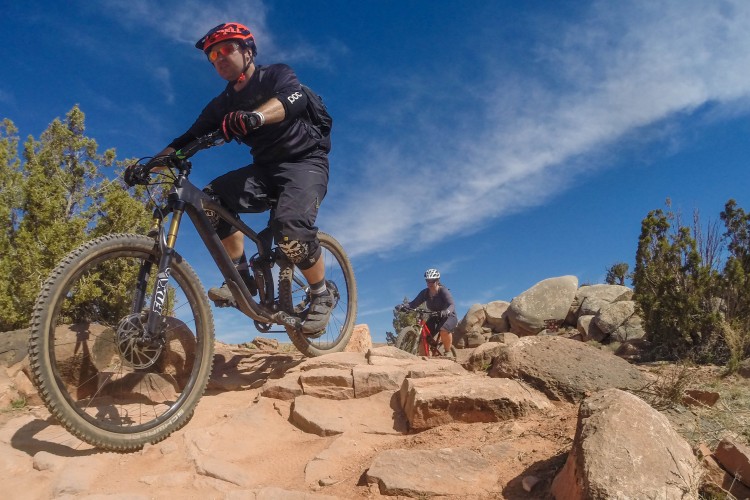 Two mountain bikers navigate a rocky trail under a clear blue sky. The first rider, wearing a black outfit and helmet, is in mid-air as they jump off a rocky section of the trail. The second rider, dressed in a gray shirt and helmet, follows closely behind. Surrounding the trail are patches of greenery and large rocks, creating a rugged outdoor environment.