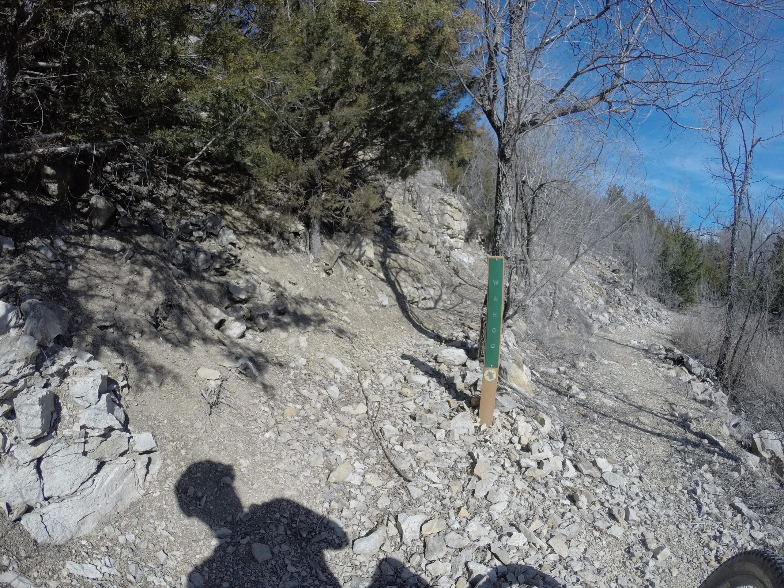 A rocky hiking trail with a green trail marker labeled "Wahoo" on the right, surrounded by trees and a clear blue sky. The shadow of a person is visible in the foreground. Melvern Riverfront Trails mountain bike trail.