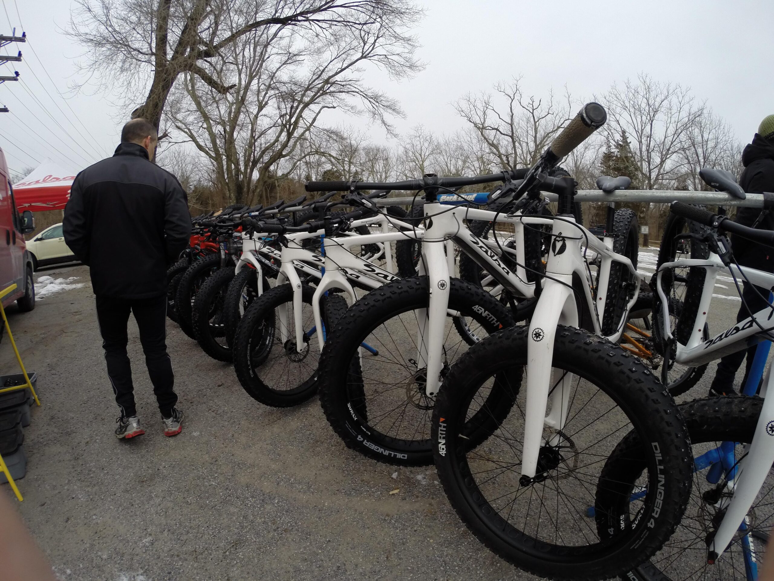 A man in a black jacket stands next to a line of white fat tire bicycles at an outdoor event, with leafless trees and a cloudy sky in the background. Several bicycles are parked on a gravel surface, showcasing their large, knobby tires. A vehicle and a tent are visible in the background. Allaire State Park mountain bike trail.