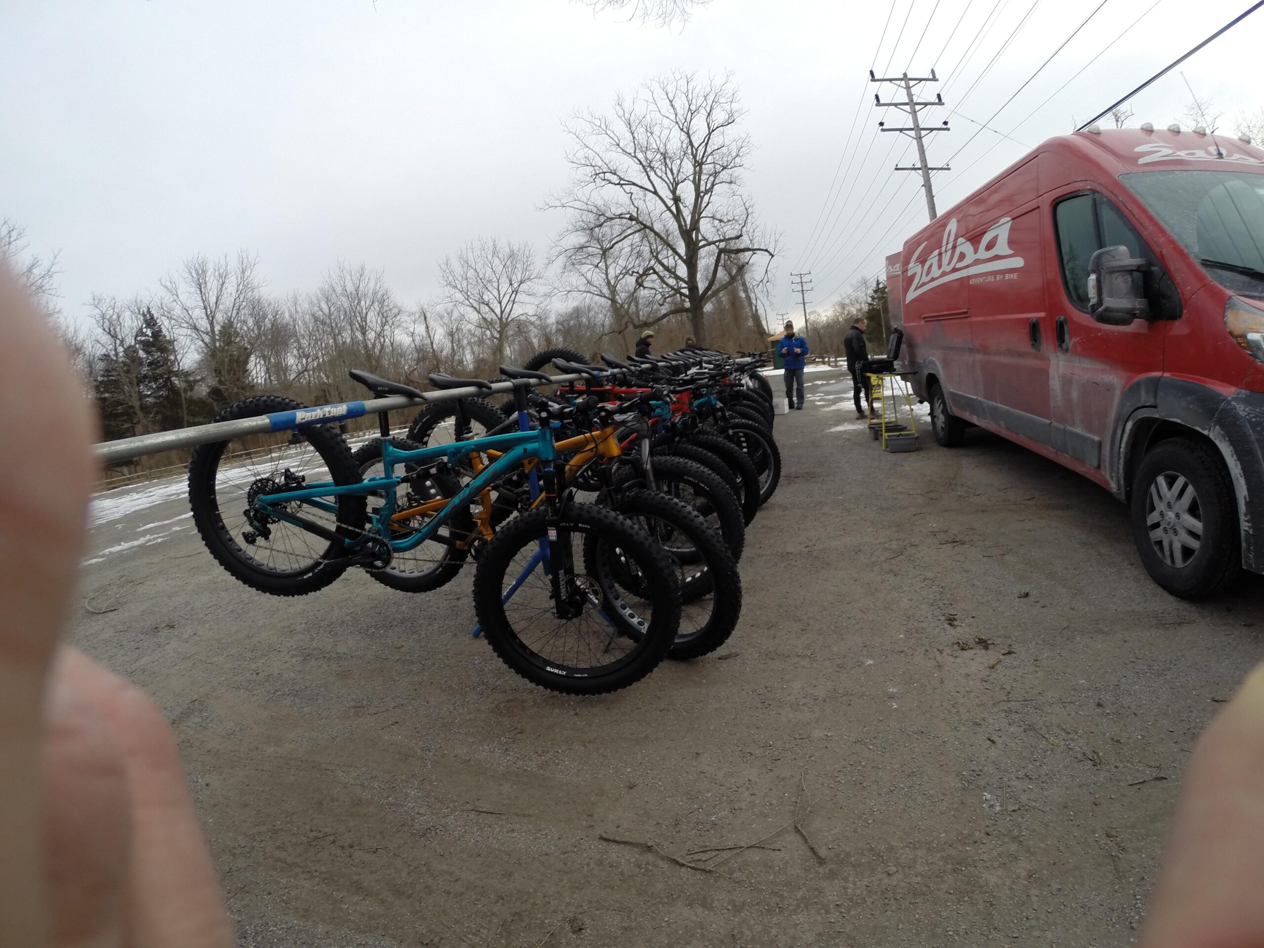 A row of colorful mountain bikes displayed on a rack next to a red van with branding. In the background, a few people are gathered near the van in a wintery landscape with bare trees and a cloudy sky. Allaire State Park mountain bike trail.