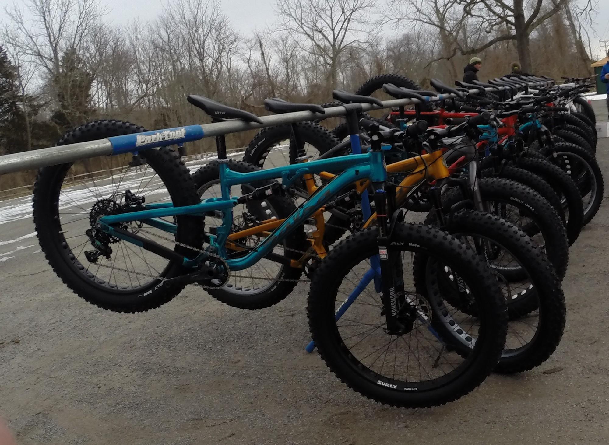 A row of mountain bikes hanging on a rack, featuring various colors including blue, orange, and red. The scene is set in a park with leafless trees in the background, and the ground shows patches of snow. The sky is overcast, indicating a cool day. Allaire State Park mountain bike trail.