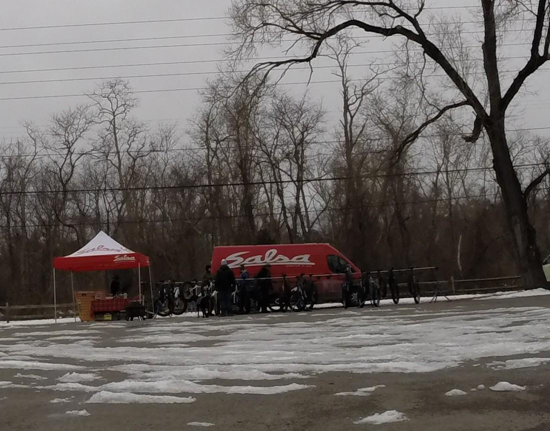 A scene featuring a red van with the logo "Salsa" parked next to a tent with the same branding. Several bicycles are displayed on racks in front of the tent, where people are gathered, and a few patches of snow are visible on the ground. Leafless trees are in the background under a cloudy sky. Allaire State Park mountain bike trail.