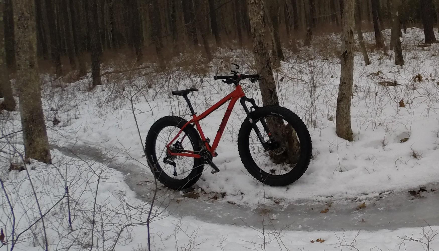 A bright red fat bike is propped against a tree in a snowy forest. The scene features a snowy trail with patches of ice and sparse vegetation, indicating winter conditions among tall trees in the background. Allaire State Park mountain bike trail.