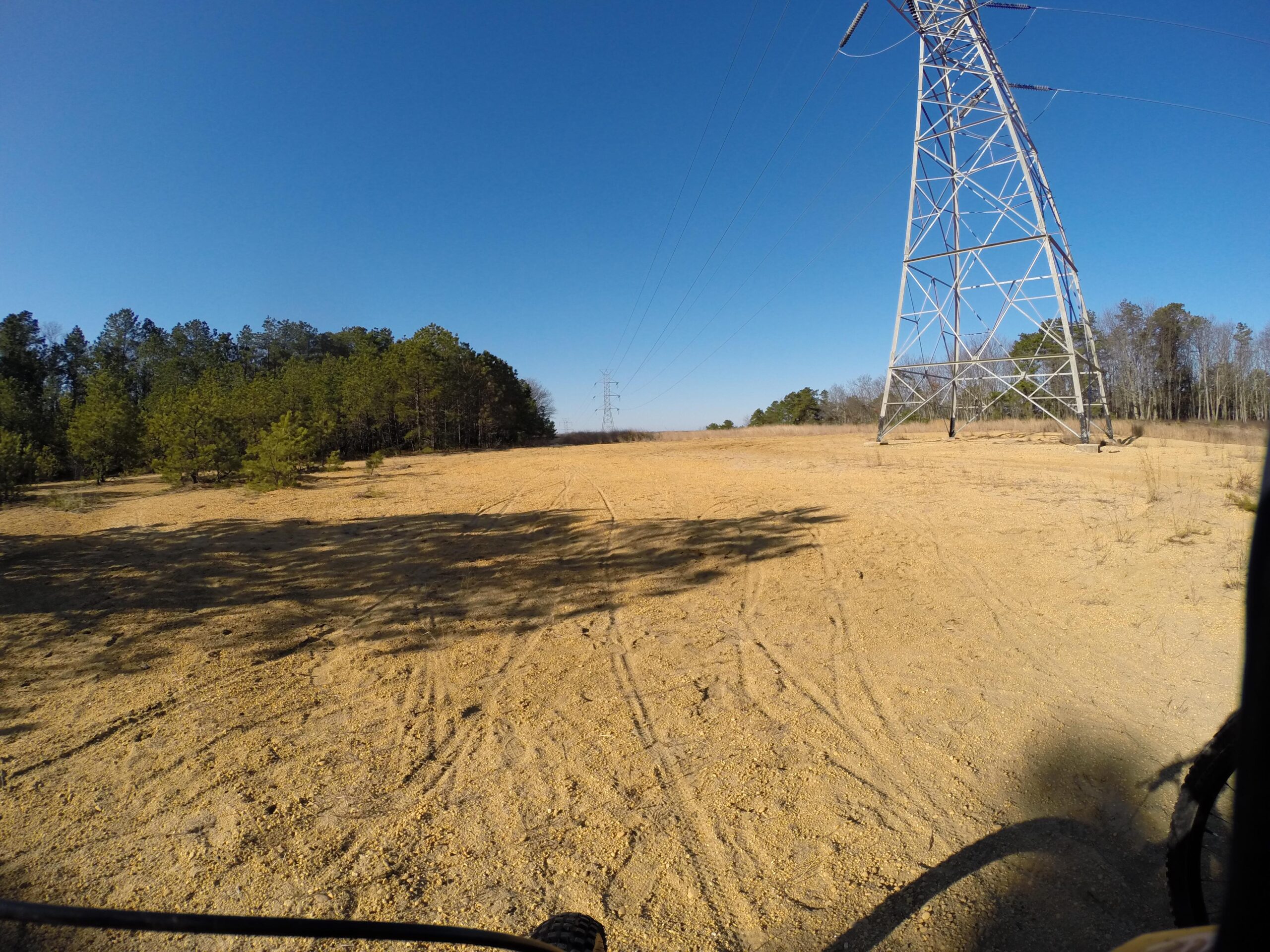 A wide view of a sandy, open field bordered by a cluster of trees on the left and a tall power transmission tower on the right, under a clear blue sky. Tire tracks are visible in the sandy area, indicating vehicular activity. Allaire State Park mountain bike trail.