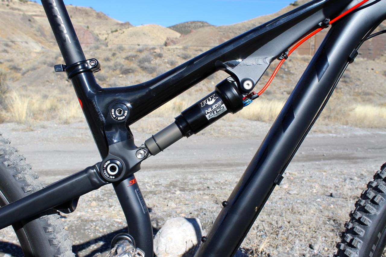 Scott Genius 930: Close-up view of a mountain bike frame featuring a rear shock absorber, with a focus on the linkage and suspension system. The image captures the bike's sleek design against a backdrop of rocky terrain and hills under a clear blue sky.