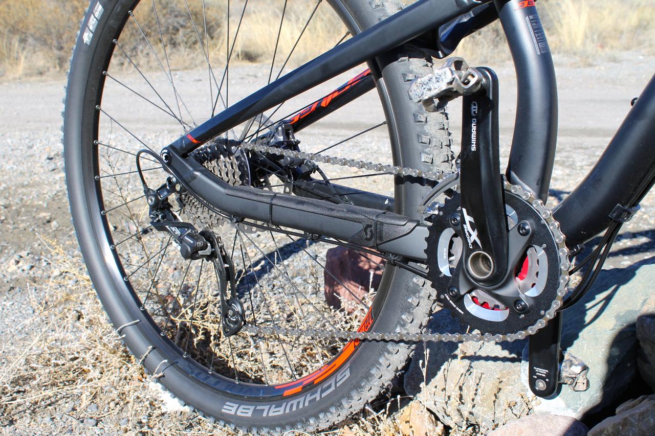 Scott Genius 930: Close-up view of a mountain bike's drivetrain, showcasing the chain, sprockets, and crankset against a backdrop of gravel and sparse vegetation.