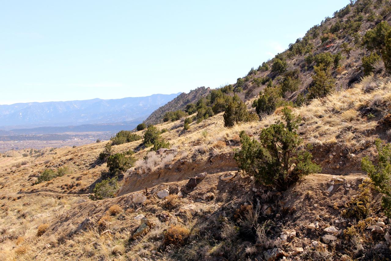 A scenic view of rolling hills and a rocky slope, covered with dry grass and sparse shrubs, against a backdrop of distant mountains under a clear blue sky. Gallineros mountain bike trail.
