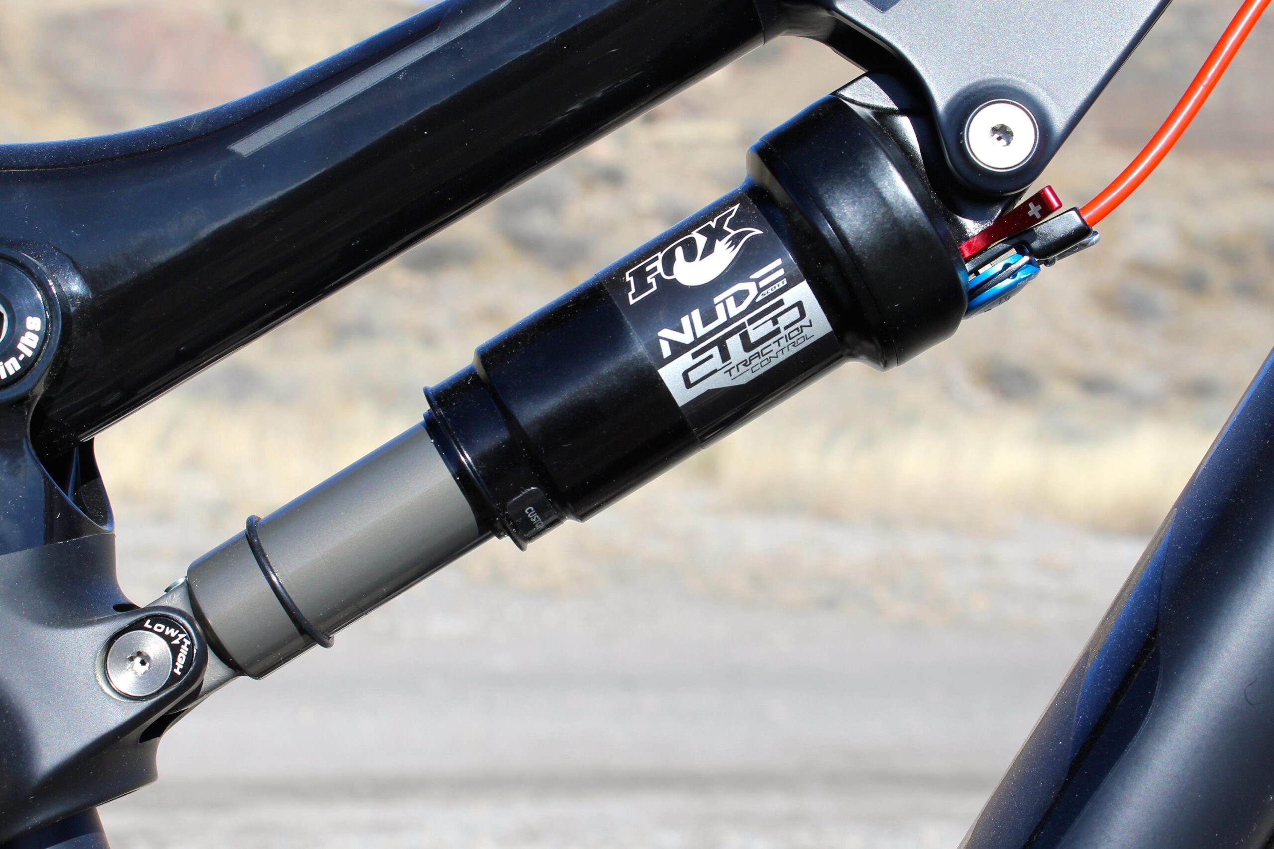 Scott Genius 930: Close-up image of a bicycle shock absorber, featuring the FOX Nude CTD model, with labels indicating settings for low and high adjustments. The shock is mounted on a dark frame, surrounded by an out-of-focus natural background.