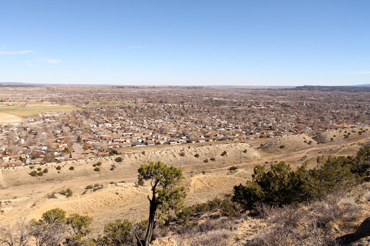 A panoramic view of a residential area set against a vast landscape, featuring scattered houses with varying roof colors, surrounded by open fields and rolling hills. The horizon extends into the distance beneath a clear blue sky, showcasing the natural beauty of the surroundings. Sparse vegetation and dirt paths are visible in the foreground. Dakota Ridge mountain bike trail.