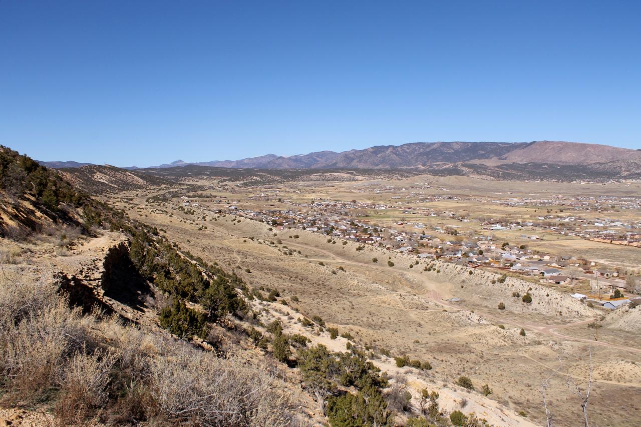 A panoramic view of a valley surrounded by mountains, featuring a small town in the foreground. The landscape showcases dry, hilly terrain with scattered vegetation and a clear blue sky above. Dakota Ridge mountain bike trail.