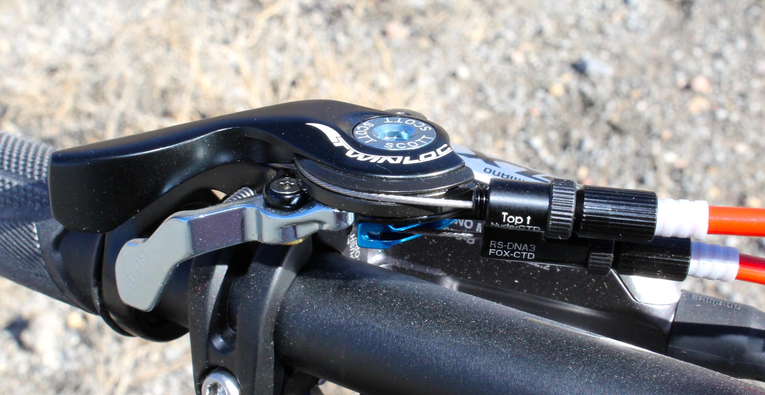 Scott Genius 930: Close-up of a bike's handlebar featuring a black TwinLoc remote control unit, which allows the rider to adjust suspension settings. The control is mounted on a dark handlebar, with visible cables connecting it to suspension components. The background shows a gravel surface, suggesting an outdoor cycling environment.